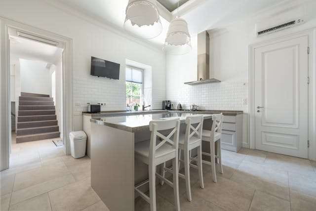 A white kitchen with a breakfast bar and white stools on a pale large-format tile floor, with a staircase visible through the doorway.