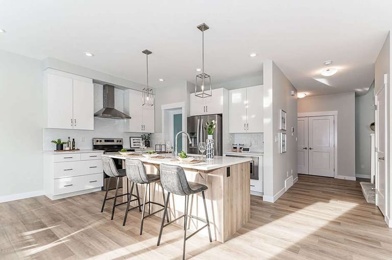Western Homes: a white kitchen with a wood-topped island and three stools, opening to a hallway with a light wood-look plank floor.