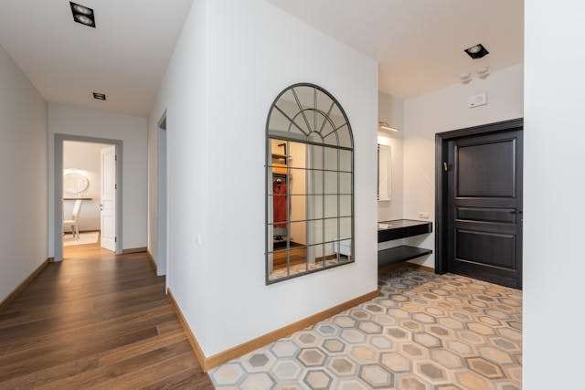 A hallway with wood plank flooring meeting a patterned hexagon tile entry, beside an arched mirror and a dark front door.