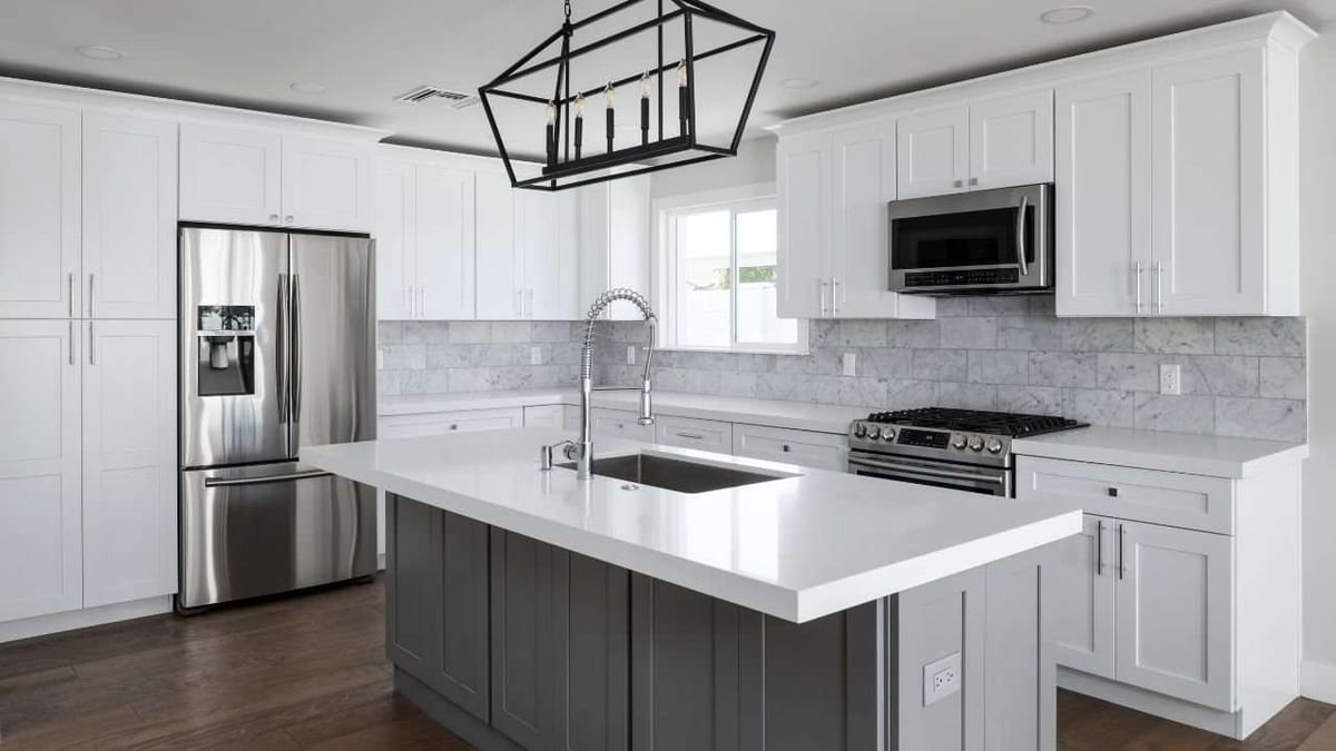 A white shaker kitchen with a grey island, white countertops and stainless appliances over a dark wood floor.