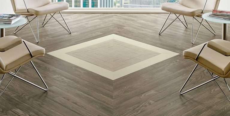 A waiting area with grey wood-look planks laid in a chevron pattern around a lighter inset square, ringed by cream lounge chairs.