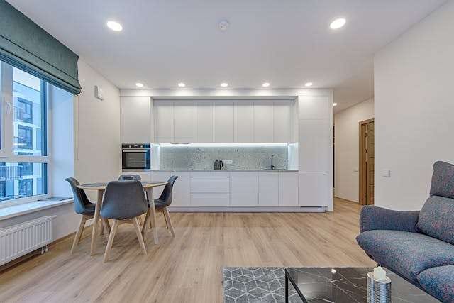 An open-plan living room and kitchen with light wood-look plank flooring, a round dining table and white cabinetry.