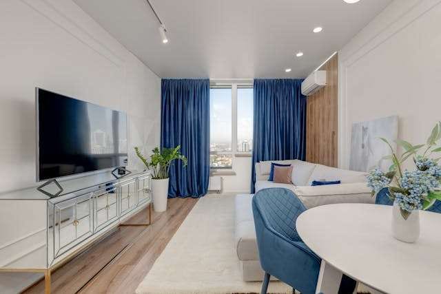 A bright living room with light wood-look flooring, a white rug, blue curtains and a white sectional sofa.
