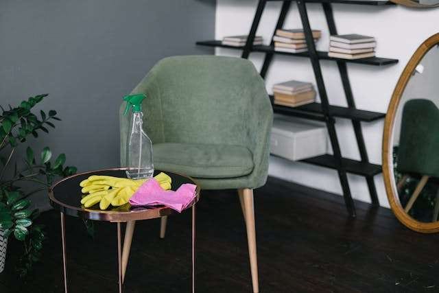 Cleaning gloves, a cloth and a spray bottle on a side table beside a green chair on a dark floor.