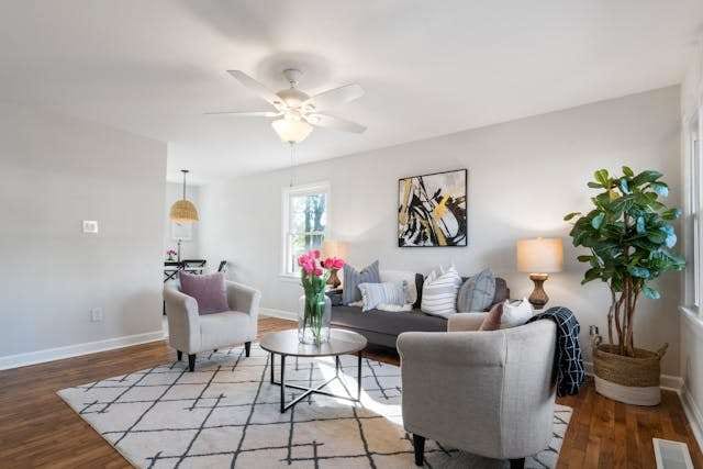 A living room with a medium-toned hardwood floor, a patterned rug, grey sofa and armchairs, and a ceiling fan.