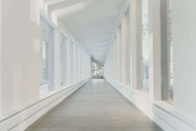 A long empty corridor with pale seamless flooring, white columns and daylight from windows along one side.