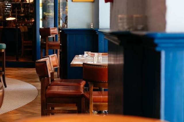 A restaurant interior with leather bar stools and small tables beside a blue-painted bar.