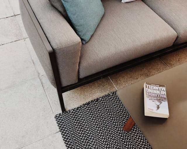 Close-up of a sofa arm and a book on a patterned rug over a pale stone tile floor.