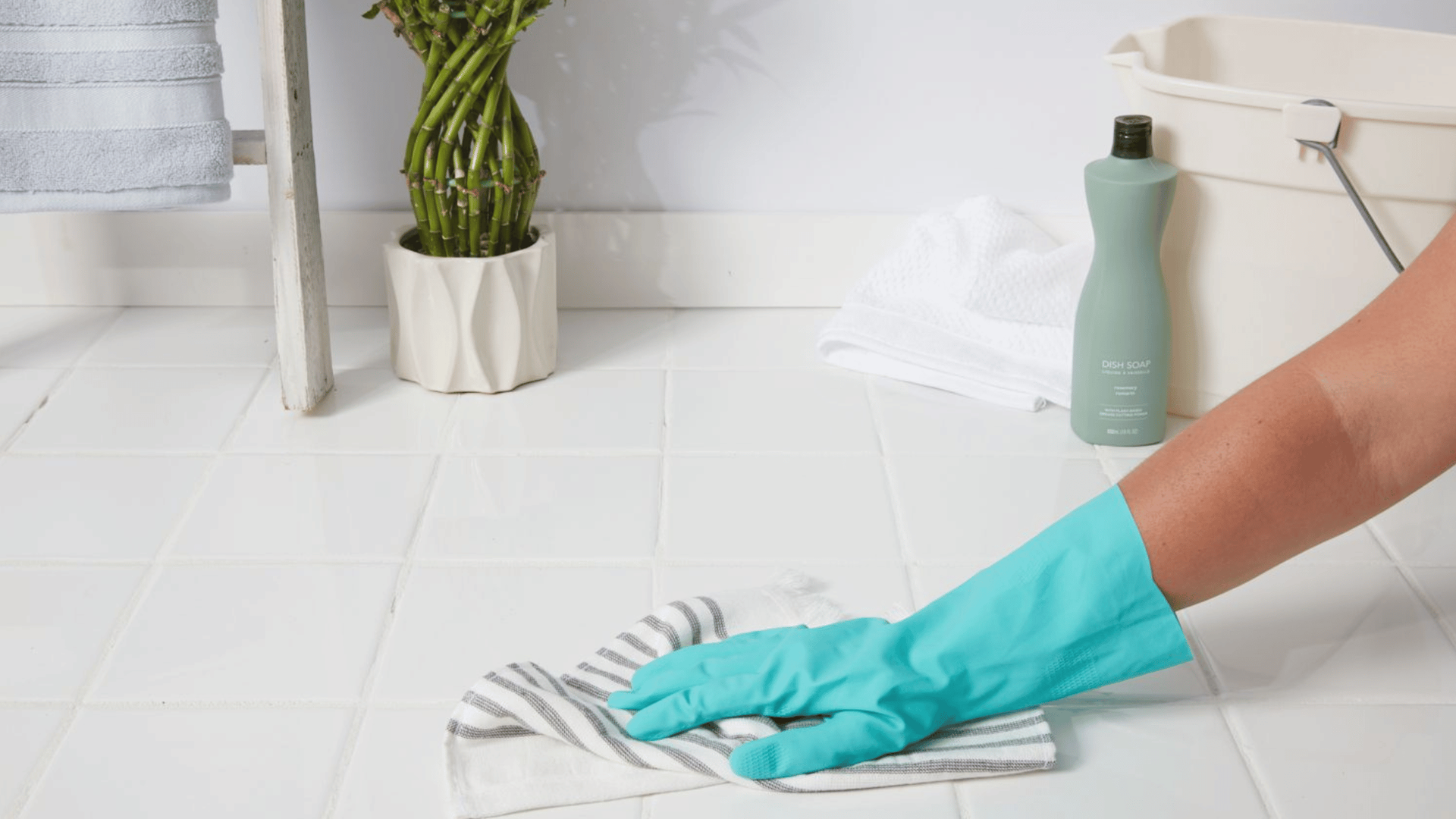 A gloved hand wiping a white tile floor with a cloth, beside a cleaning bottle and a potted plant.