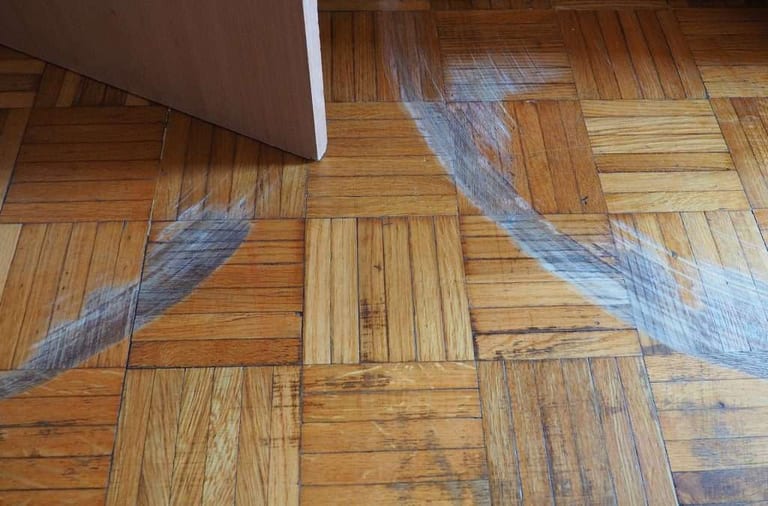 A furniture leg dragging across a wood parquet floor, with blue arrows tracing the scratches left behind.