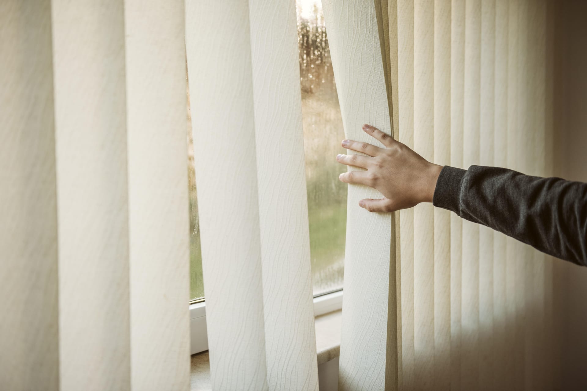 A hand drawing back cream vertical blinds at a window.