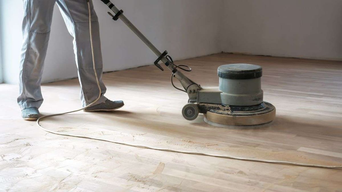 A worker running a floor buffer over a sanded, unfinished wood floor.