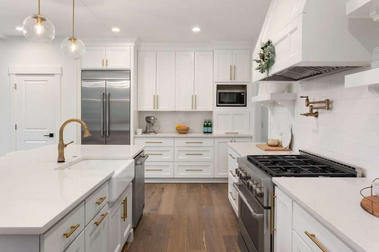 A white kitchen with brass fixtures, a farmhouse sink, quartz counters and a medium-brown wood floor.