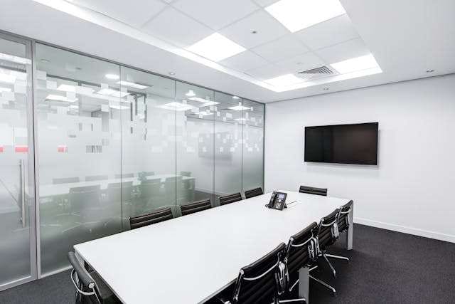A boardroom with a white table, black chairs and frosted glass walls over dark carpet tile.