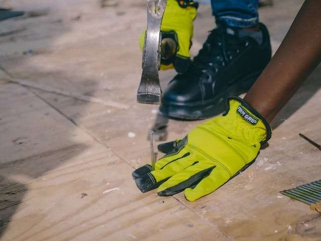 Gloved hands using a hammer and pry bar on a plywood subfloor.