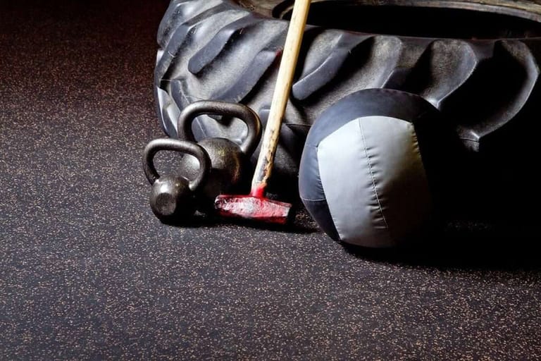 Kettlebells, medicine balls and a barbell resting on dark speckled rubber gym flooring.
