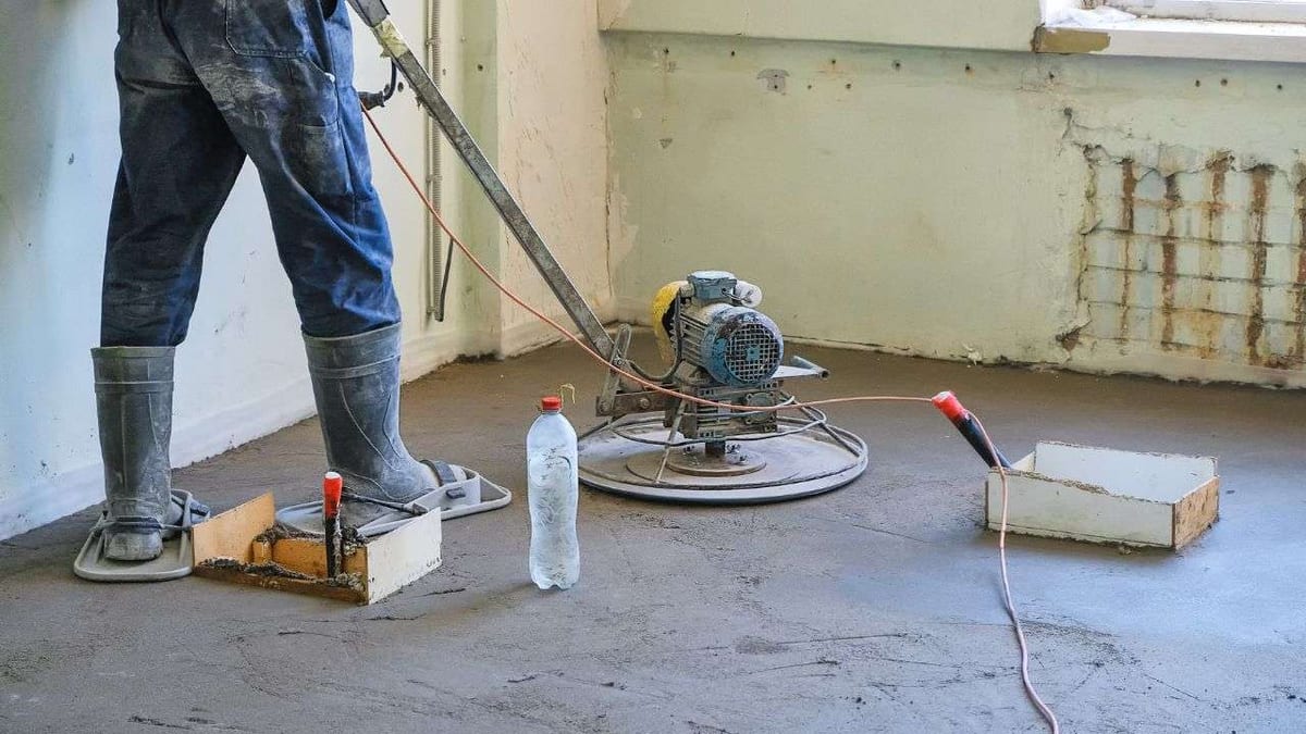 A worker smoothing a concrete subfloor with a power trowel in a room under renovation.