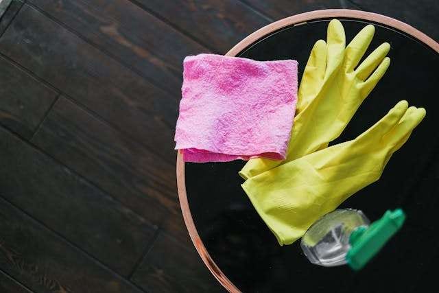 Yellow rubber gloves, a pink cloth and a cleaning bottle on a small table over a dark wood floor.