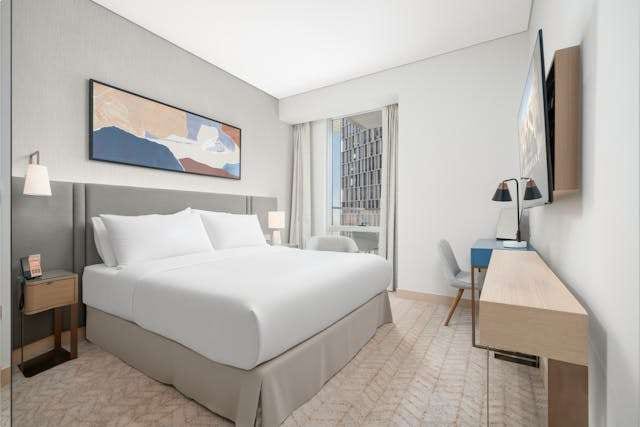 A hotel-style bedroom with a white bed, a wooden desk and pale patterned carpet.