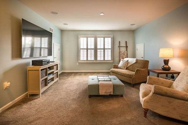 A living room with beige carpet, upholstered armchairs and an ottoman under plantation-shuttered windows.