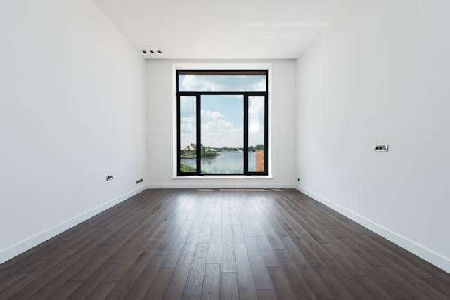 An empty white room with a dark wood plank floor and a tall black-framed window.