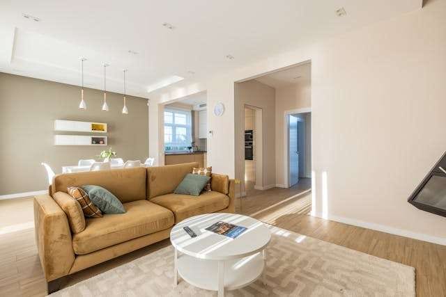 A living room with light wood flooring, a tan sofa and a white round coffee table, with a dining area beyond.