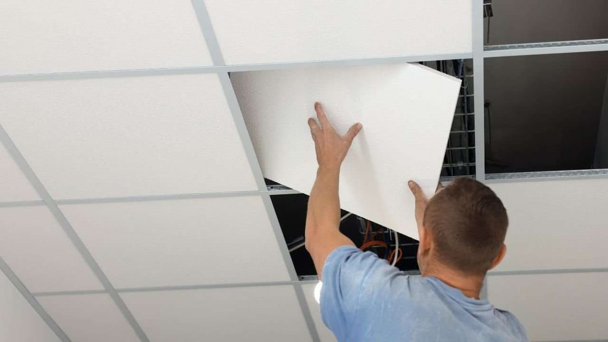 A man lifting a white panel into a suspended ceiling grid.