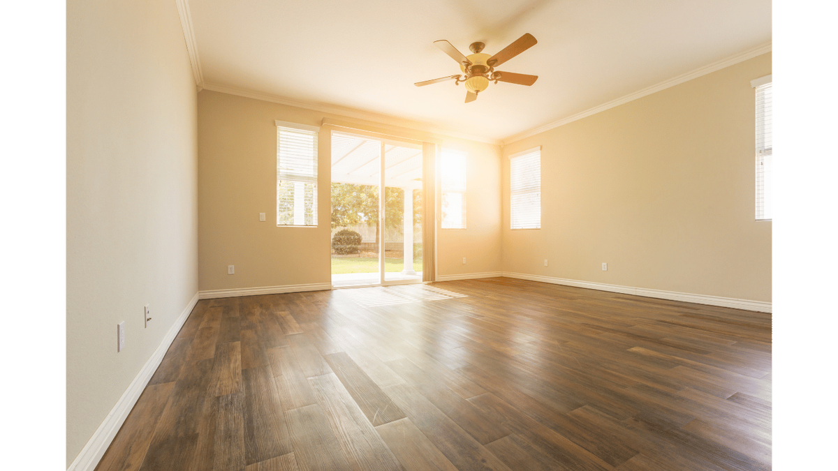 Dark brown wood-look plank flooring in an empty sunlit room with a ceiling fan and sliding patio doors.