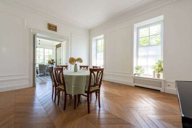 A dining room with a medium-brown herringbone wood floor, white panelled walls and a round table.
