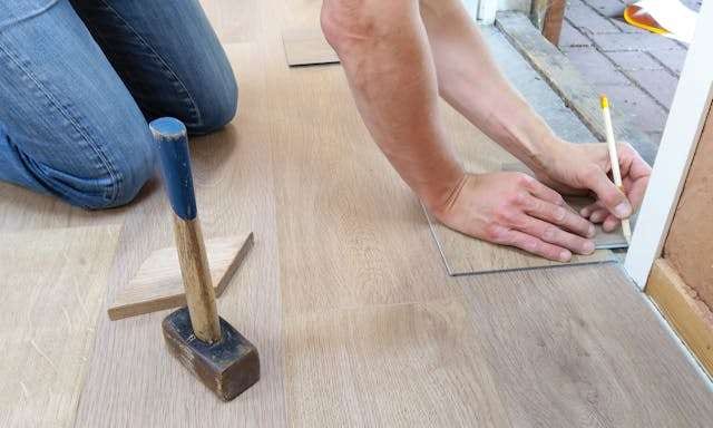 An installer marking a light wood-look plank with a pencil, a mallet beside him on the floor.