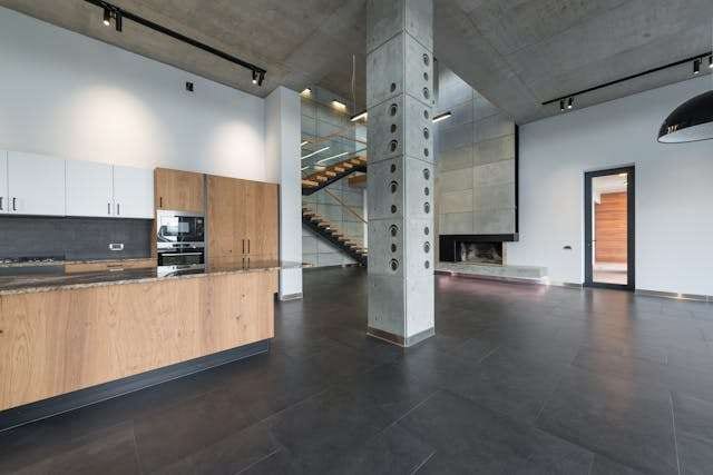 An open-plan kitchen with wood cabinetry, concrete columns and dark large-format tile flooring.
