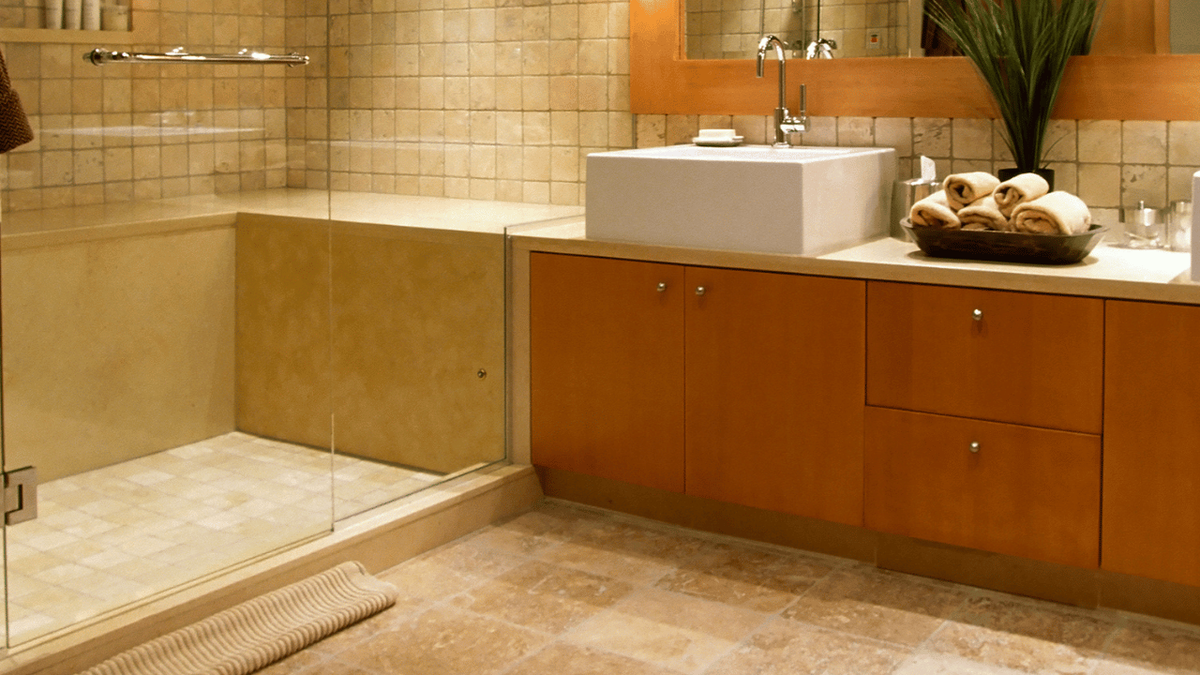 Beige stone-look tile flooring in a bathroom with a walk-in glass shower, orange wood vanity and vessel sink.