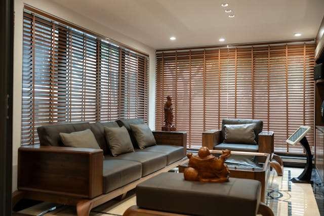 A living room with wood venetian blinds across tall windows, a dark leather sofa and wooden furniture.
