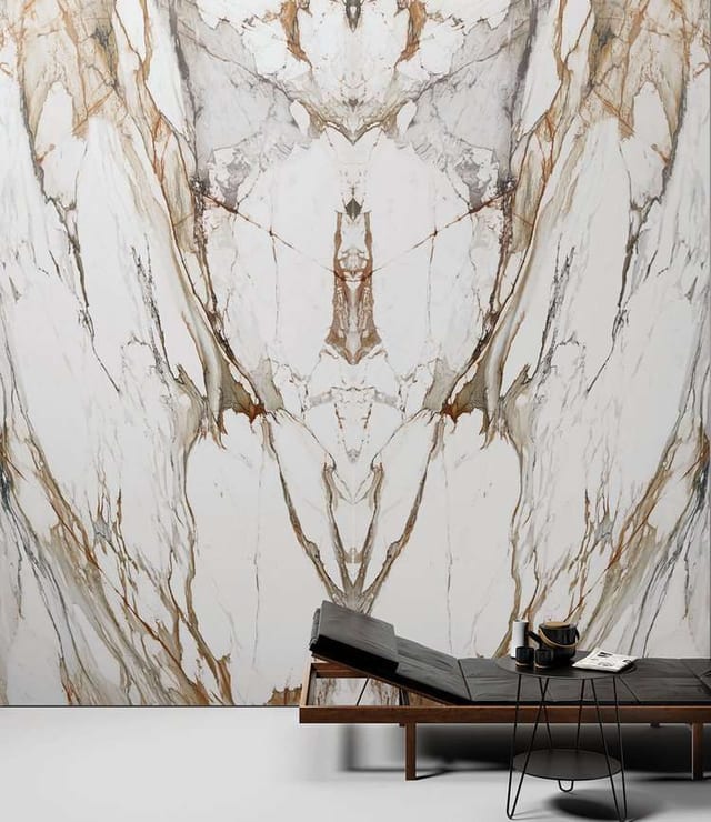 A floor-to-ceiling white marble-look porcelain slab with bookmatched gold and grey veining behind a lounge chair.