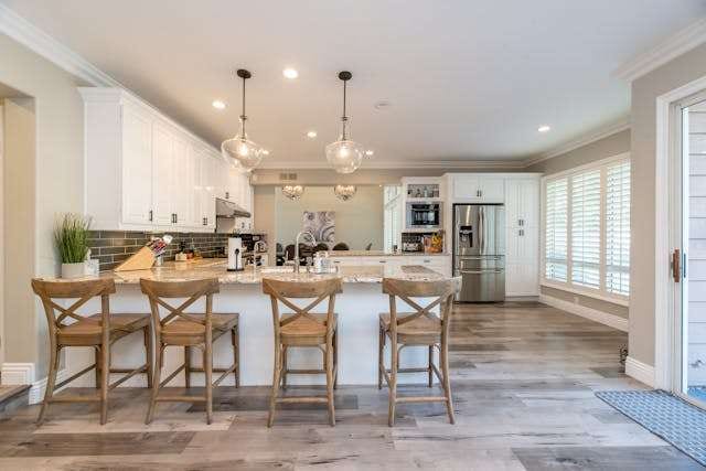 A white kitchen with a large island, wooden bar stools and grey wood-look plank flooring.