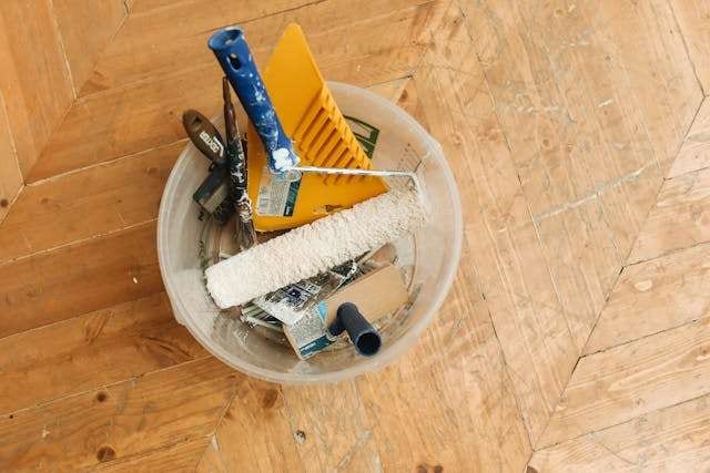 A bucket of painting and flooring tools sitting on a worn pine plank floor.