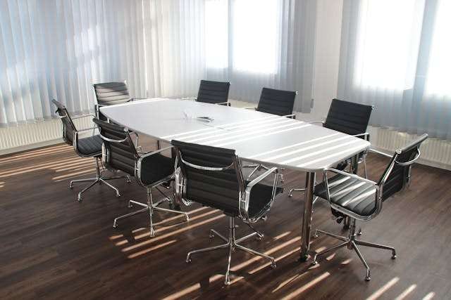 A boardroom with a white table, black office chairs and a dark wood-look floor in striped sunlight.