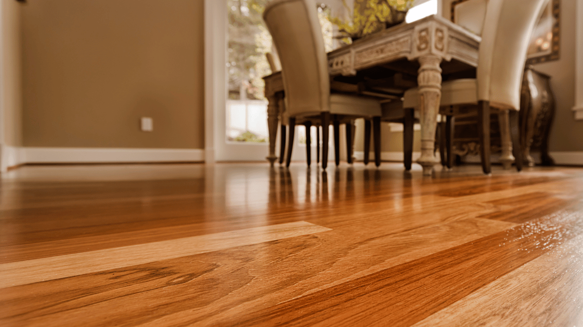 Glossy golden hardwood strip flooring in a dining room, with a carved table and upholstered chairs in the background.
