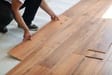 An installer laying warm brown wood planks with a visible tongue-and-groove edge.