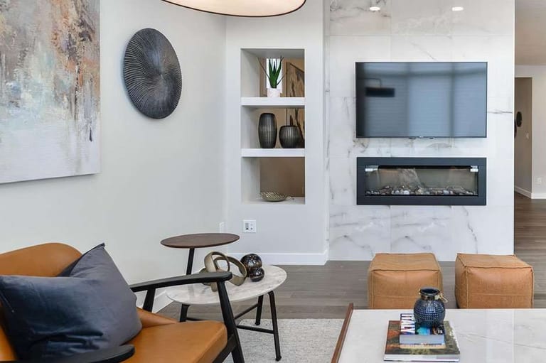 Modern Vibes staged home: a living room with a marble-look fireplace surround, built-in niches, tan leather ottomans and a pale rug.