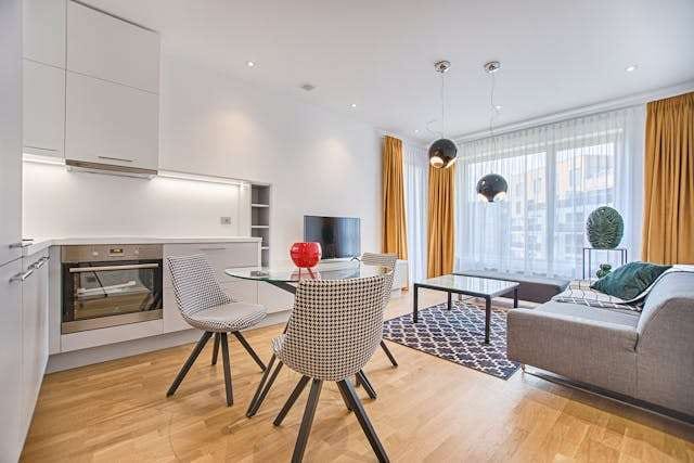 An open-plan kitchen and living room with light oak-look flooring, a glass dining table and a grey sofa.