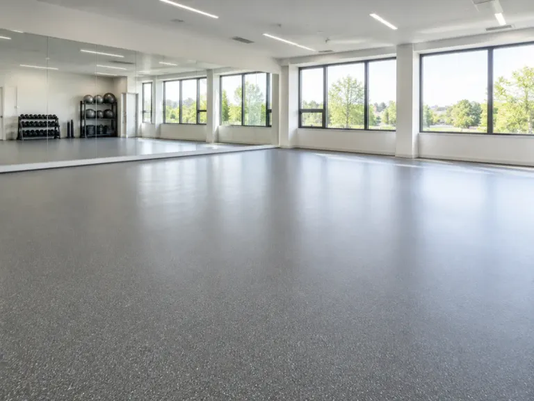 An empty fitness studio with continuous grey speckled roll-out rubber flooring, a mirror wall and large windows.