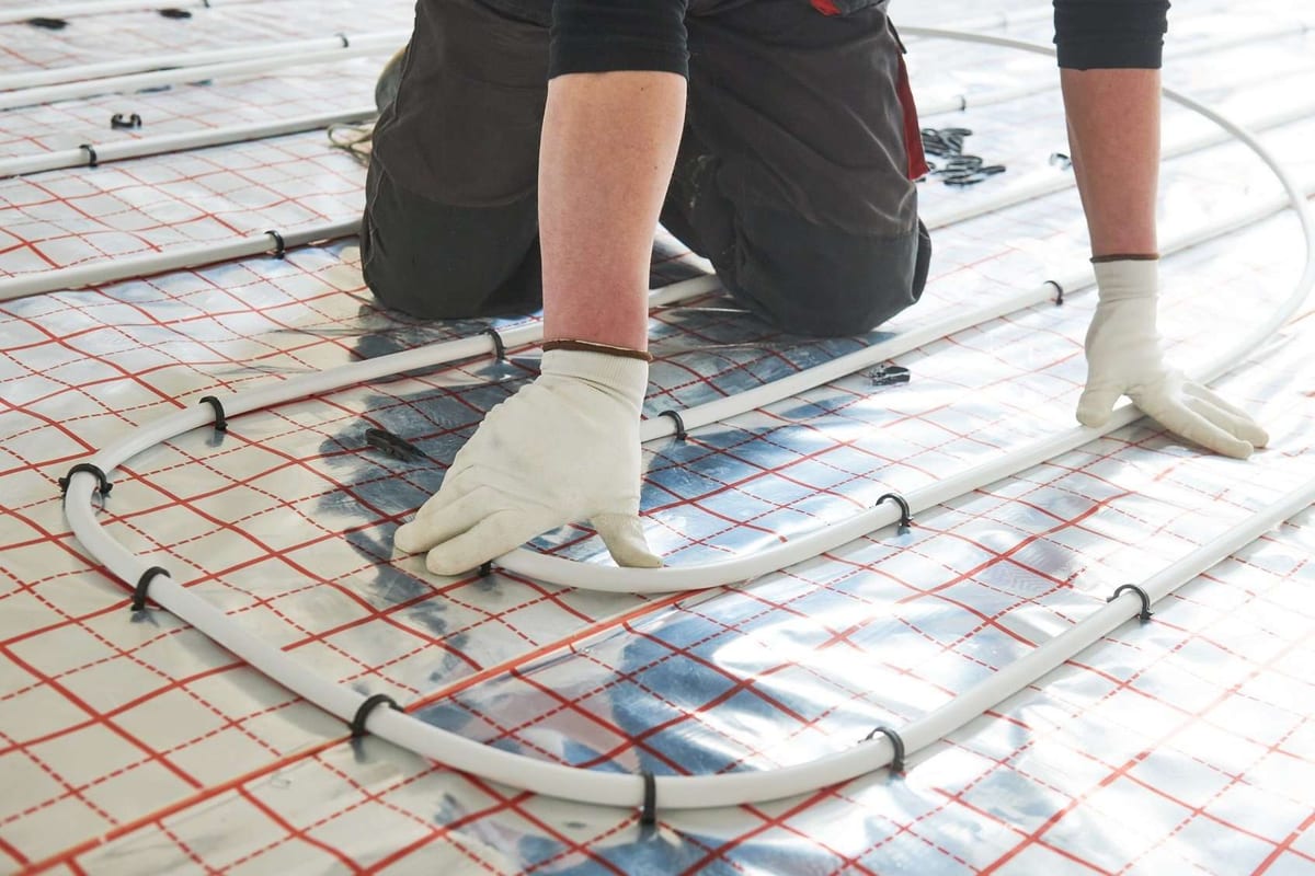 Gloved hands laying white in-floor heating pipe in loops over a gridded insulation mat.
