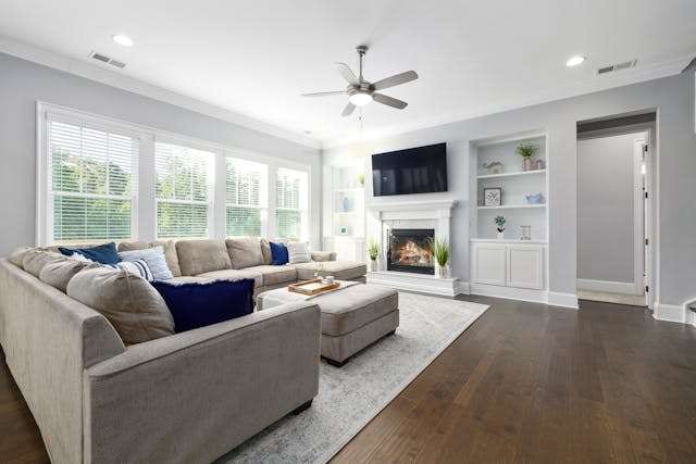A family room with a dark wood-look floor, a grey sectional, a fireplace and built-in shelves.