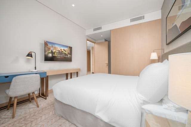 A hotel-style bedroom with a white bed, a slim desk and pale patterned carpet.