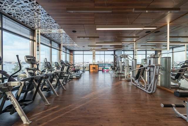 A fitness centre with treadmills and weight machines on a dark wood-look floor beside tall windows.