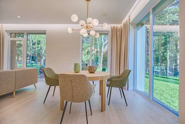 A dining area with a round wooden table, green chairs and a pale wood floor beside large windows.
