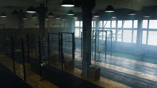 A dim industrial gym with squat racks on rubber flooring beside large windows.