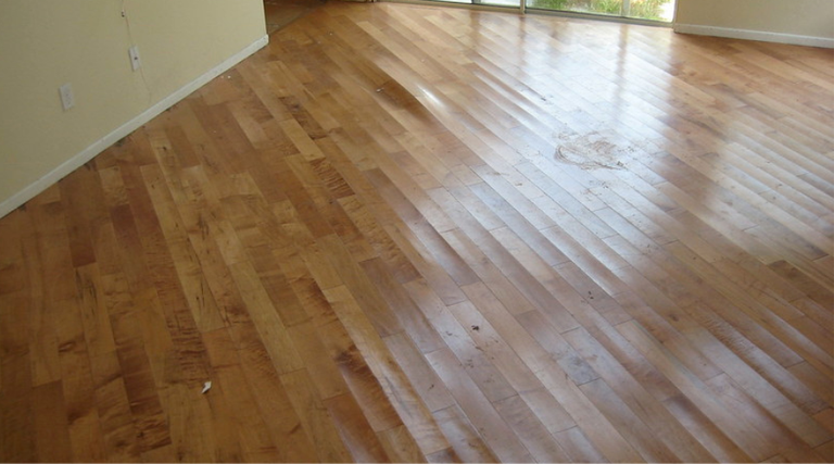 Worn honey-toned hardwood flooring in an empty room, with dull cloudy patches in the finish.