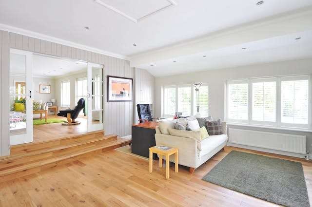 A sunlit living room with a raised step and light wood plank flooring, a cream sofa and a grey rug.